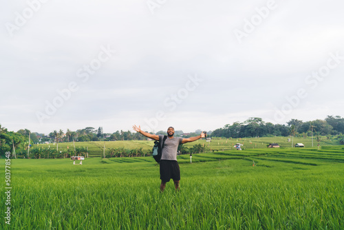 Portrait of cheerful male photographer with touristic backpack and waterproof GOPRO camera technology raising hands and smiling at camera while feeling satisfied at rice fields, getaway traveling