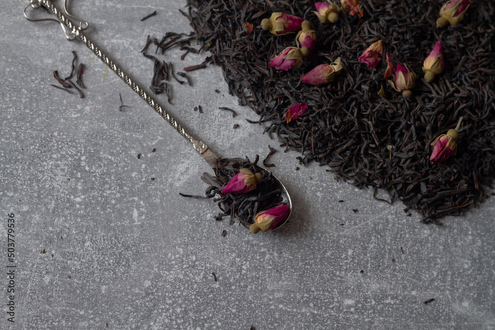 Black leaf tea with dry pink rose buds on a spoon on a gray background