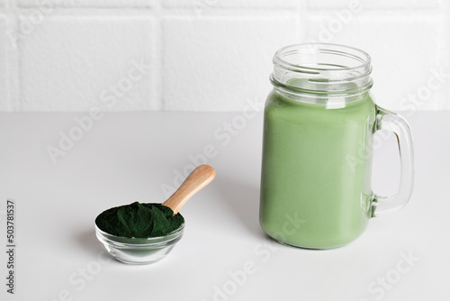 fresh green smoothie in glass bottle and spirulina powder with spoon on wooden background. useful habits, self care and healthy lifestyle. copy space