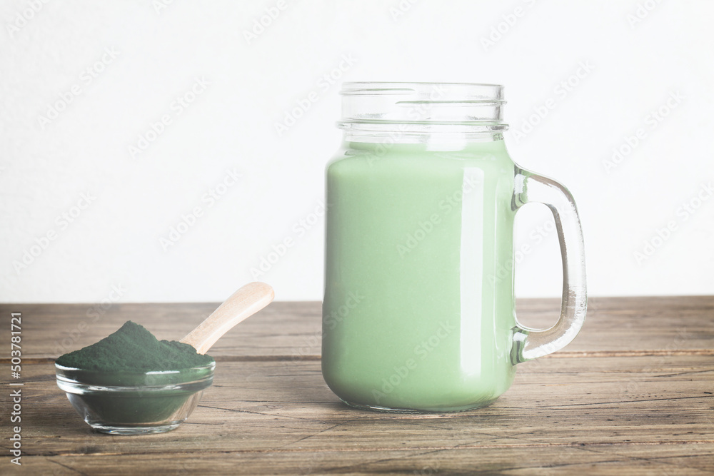 fresh green smoothie in glass bottle and spirulina powder with spoon on wooden background. useful habits, self care and healthy lifestyle. copy space