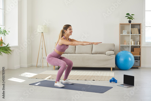 Happy woman having training workout at home with her virtual fitness instructor. Sporty young Caucasian lady standing on yoga mat in living room and doing squat exercise near laptop with empty screen