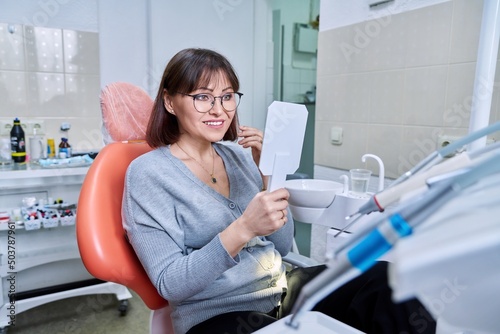 Smiling middle aged woman in dental chair with mirror looking at her teeth