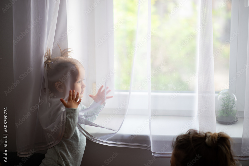 baby girl behind curtain holding hands up, mother is looking, natural home window background - obrazy, fototapety, plakaty 