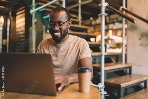 Cheerful copywriter in eyewear checking received email messages on laptop computer using coworking internet for doing distance job, African American programmer in spectacles chatting and smiling