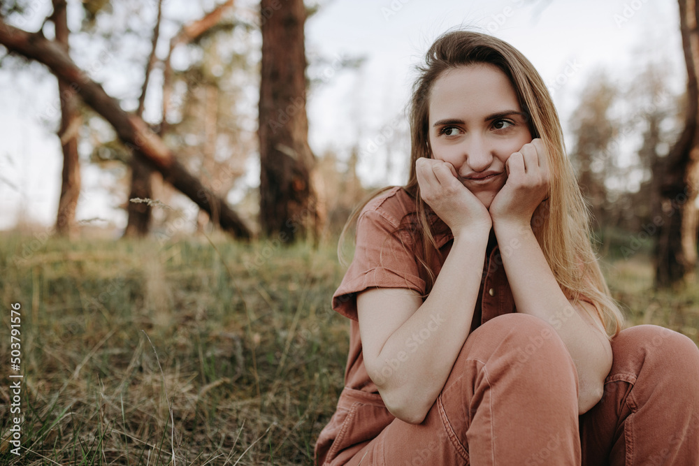Funny blonde girl with grumpy face putting her hands on her face and sitting in forest. Female model with comical and ridiculous face Funny blonde girl with grumpy face putting her hands on her face and sitting in forest. Female model with comical and ridiculous face