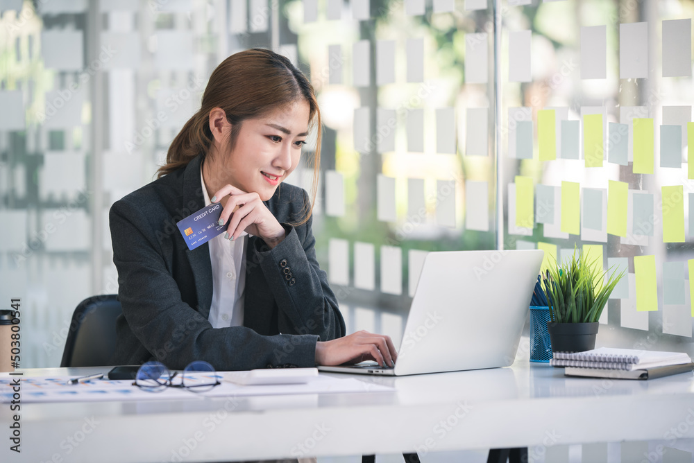 Asian woman holding a credit card and using a laptop computer Businesswoman working at modern office, online shopping, ecommerce, internet banking, spending money, credit card working concept. Asian woman holding a credit card and using a laptop computer Businesswoman working at modern office, online shopping, ecommerce, internet banking, spending money, credit card working concept.
