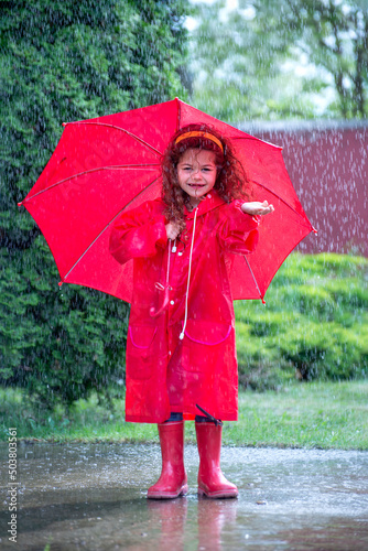 Happy funny child with red umbrella under shower.Girl is wearing red raincoat and enjoying rainfall. Kid playing on the nature outdoors.
