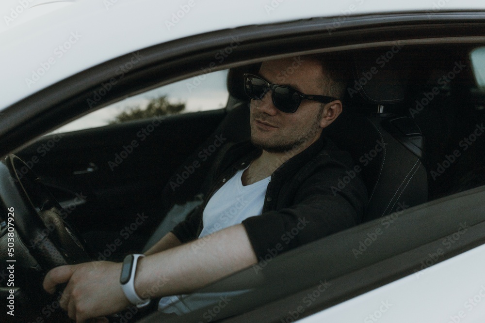 Handsome man driver with black sunglasses in black T-shirt and white shirt sits in modern white sport car and looks at the sunset outdoors. Fresh weekend out of city near riverside. Dark tone photo
