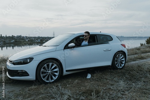 Handsome man in black T-shirt and white shirt with jeans and sneakers poses near modern white sport car and looks at the sunset outdoors. Fresh weekend out of the city near riverside. Dark tone photo 