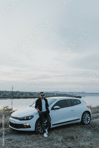 Handsome man in black T-shirt and white shirt with jeans and sneakers poses near modern white sport car and looks at the sunset outdoors. Fresh weekend out of the city near riverside. Dark tone photo 