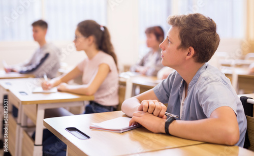 Portrait of positive teenage male pupil sitting at desk studying in classroom