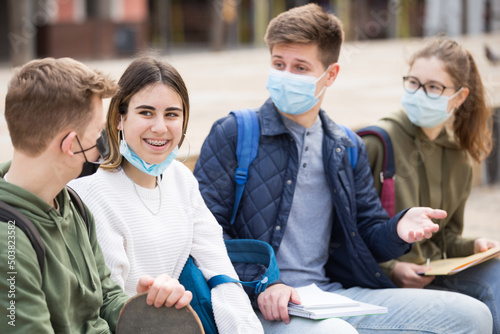 Cheerful teenagers wearing face masks for disease protection sitting together, talking and having fun outdoors