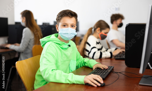 Diligent tween boy in protective face mask studying with classmates in modern computer lab at school during coronavirus pandemic. Concept of compelled precautions
