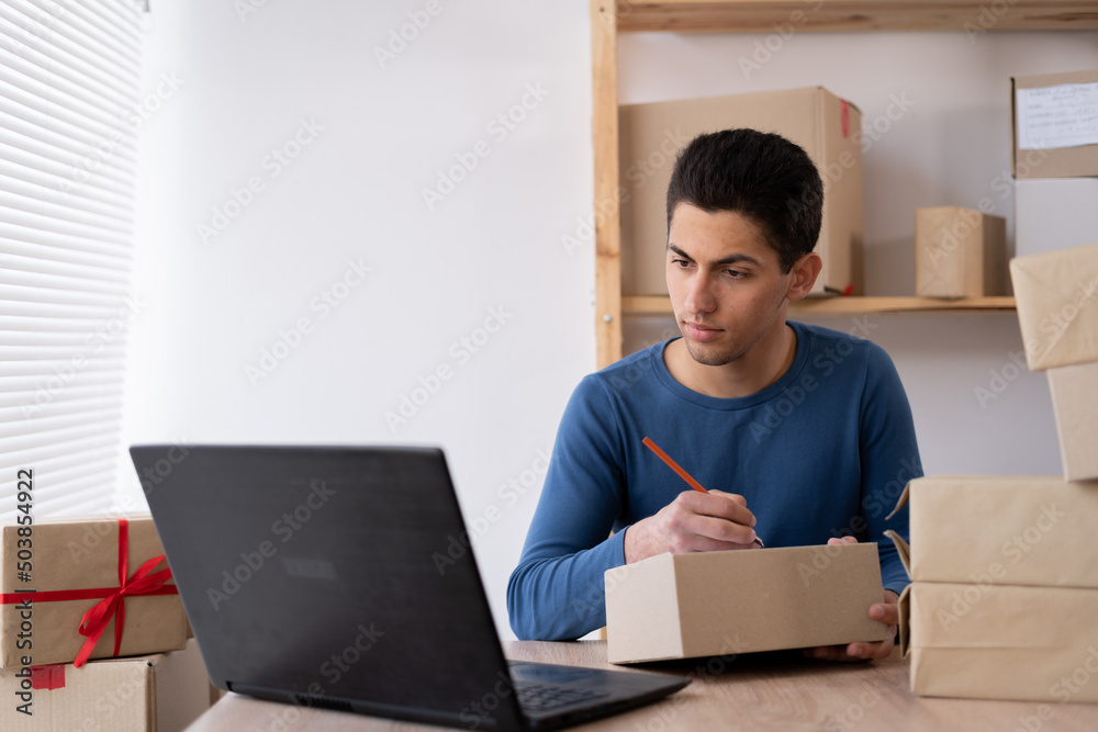 a small business startup owner working at home with laptop. Online sale. Arabic man sits at a table against the backdrop of a rack with boxes and writes the delivery address a small business startup owner working at home with laptop. Online sale. Arabic man sits at a table against the backdrop of a rack with boxes and writes the delivery address