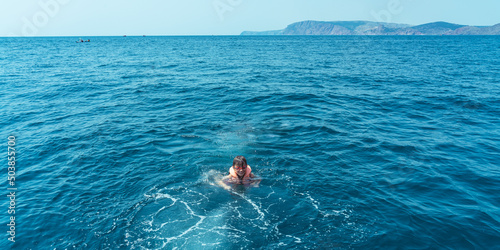 Happy caucasian boy 8 years old in a life jacket and goggles swims in the sea near the boat looking at the photographer. The concept of a healthy lifestyle, doing sports