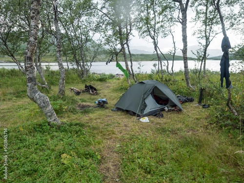 Small open green tent with backpack and hiking gear on grassy shore of Tarra river with birch trees. Swedish Lapland Landscape with green hills at Padjelantaleden hiking trail. Summer day