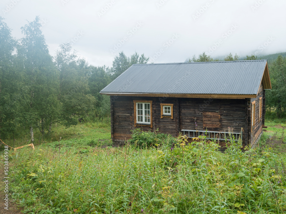 Fototapeta premium View of STF Njunjes Mountain cabin on a meadow, moody rainy day with thick fog. Wooden cottage lies on the banks of the Tarra river, at Padjelantaleden hiking trail. Lapland summer landscape