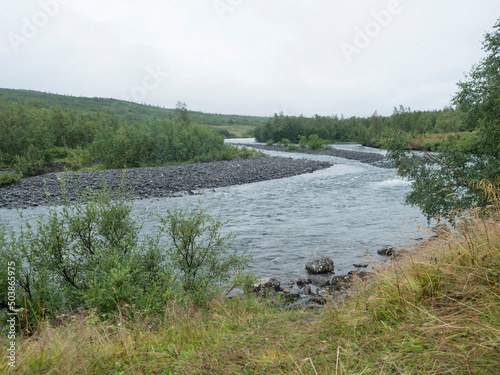 View of winding Tarra river with birch bush, gravel, grass and granite rock. Northern landscape in Swedish Lapland at Padjelantaleden hiking trail. Moody, rainy day, cloudy sky, summer. photo