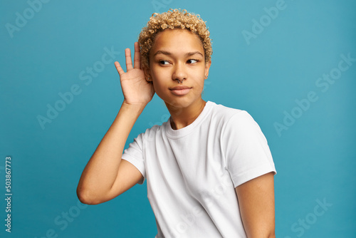 Good-looking curious dark-skinned female in white casual clothes putting hand over ear to hear better, listening to gossip or secret, having blonde curls, nose piercing and earrings, isolated on blue photo