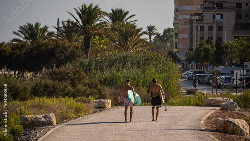 Fotografiet Back view of two surfers walking along the boulevard with surfboards