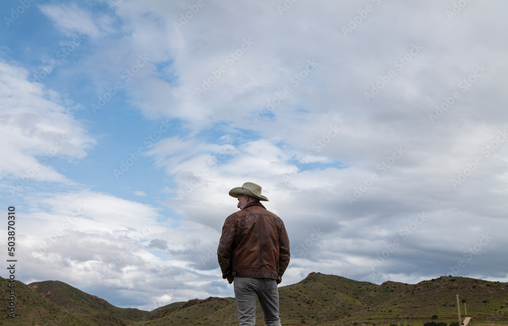 Rear view of adult man in cowboy hat on field against cloudy sky