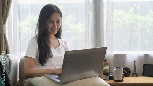 Pretty young asian woman resting on couch and using laptop, surfing internet, checking email.