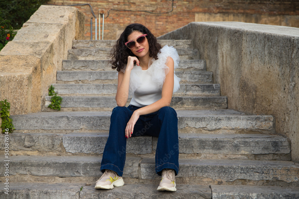 beautiful young woman with dark, curly hair sitting on a staircase in a park. The woman wears sunglasses and makes different expressions. Expressions and lifestyle concept