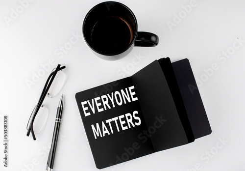 Everyone matters written text in small black notebook with coffee , pen and glasess on white background. Black-white style