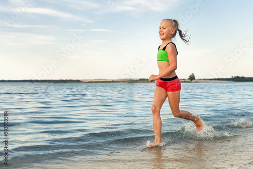 Cute adorable sporty caucasian blond school girl enjoy having fun running water by lake or sea sand beach splashing against blue sky on summer day. Summertime children healthy vacation concept