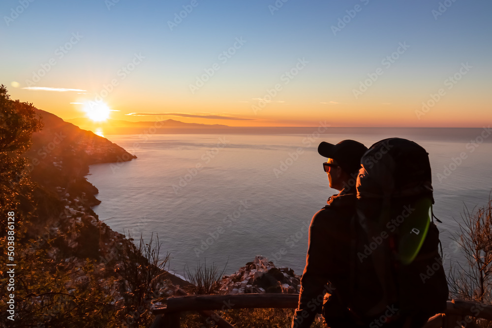 Active man with backpack watching sunrise from hiking trail Path of Gods between Positano and Praiano, Amalfi Coast, Campania, Italy, Europe. Calm water surface reflecting sun ray at Mediterranean Sea