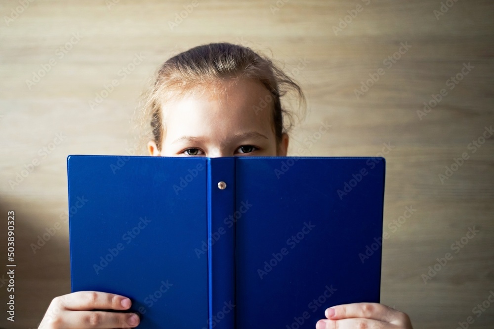 little girl child reads a blue book and looks out from behind it, eyes and a smile are visible, school tomorrow little girl child reads a blue book and looks out from behind it, eyes and a smile are visible, school tomorrow