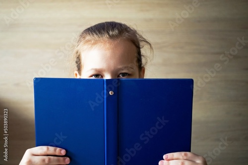 little girl child reads a blue book and looks out from behind it, eyes and a smile are visible, school tomorrow