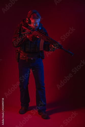 soldier in full gear with weapons. a man in headphones, body armor, with a backpack and a belt. red background. colored, blue-red light © Ольга Новицкая