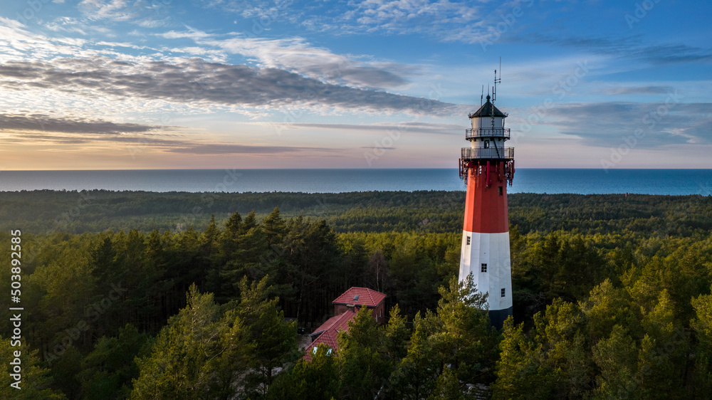 Aerial view of the lighthouse surrounded by the lush green forest. Sea in the distance. Blue sky and pastel clouds. Panoramic view.