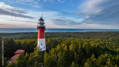 Aerial view of the lighthouse surrounded by the lush green forest. Sea in the distance. Blue sky and pastel clouds. Panoramic view.