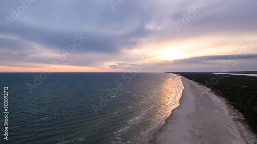 Landscape of the sandy beach during cloudy evening. Aerial view of the pine forest. Rays of the sun on the water.