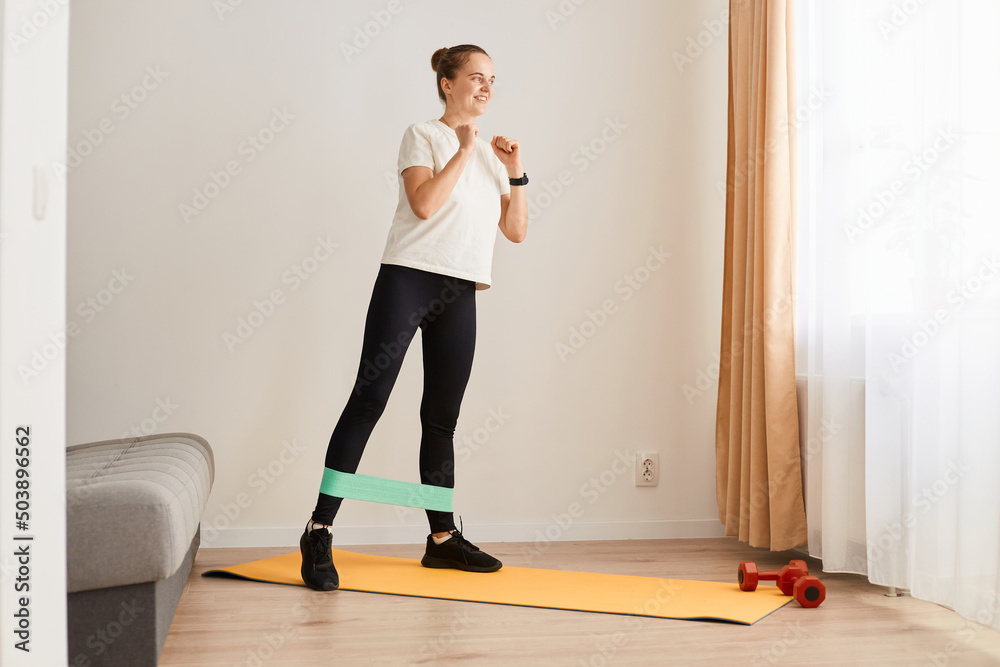 Full and lower body workout. Sporty beautiful woman exercising with elastic rubber band lifting and kicking leg aside standing on yoga mat, Bodyweight training