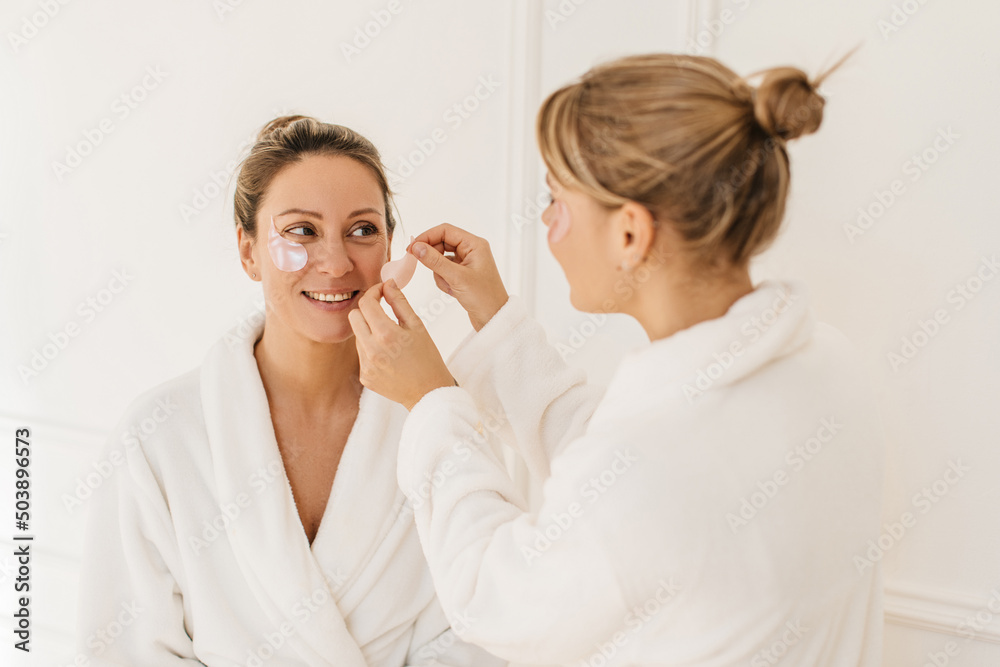 Caucasian blonde girl puts patches on face of adult woman sitting indoors. Ladies wear white terry robes. Health care concept Caucasian blonde girl puts patches on face of adult woman sitting indoors. Ladies wear white terry robes. Health care concept