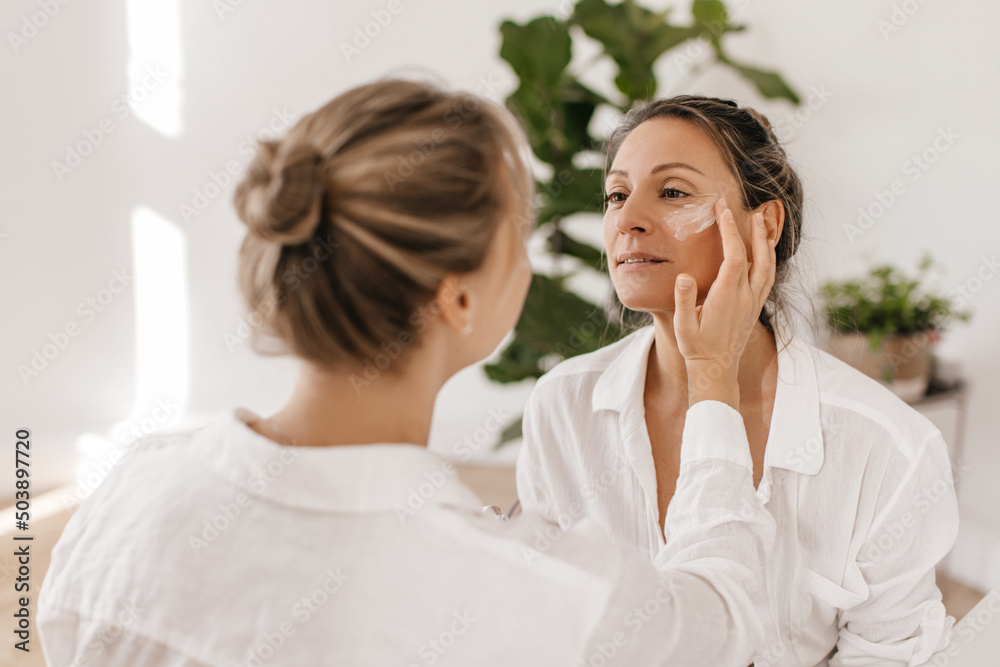 Two modern caucasian women are enjoying professional cosmetic product indoors. Blondes with collected hair in white shirts. Skin care and hydration concept Two modern caucasian women are enjoying professional cosmetic product indoors. Blondes with collected hair in white shirts. Skin care and hydration concept