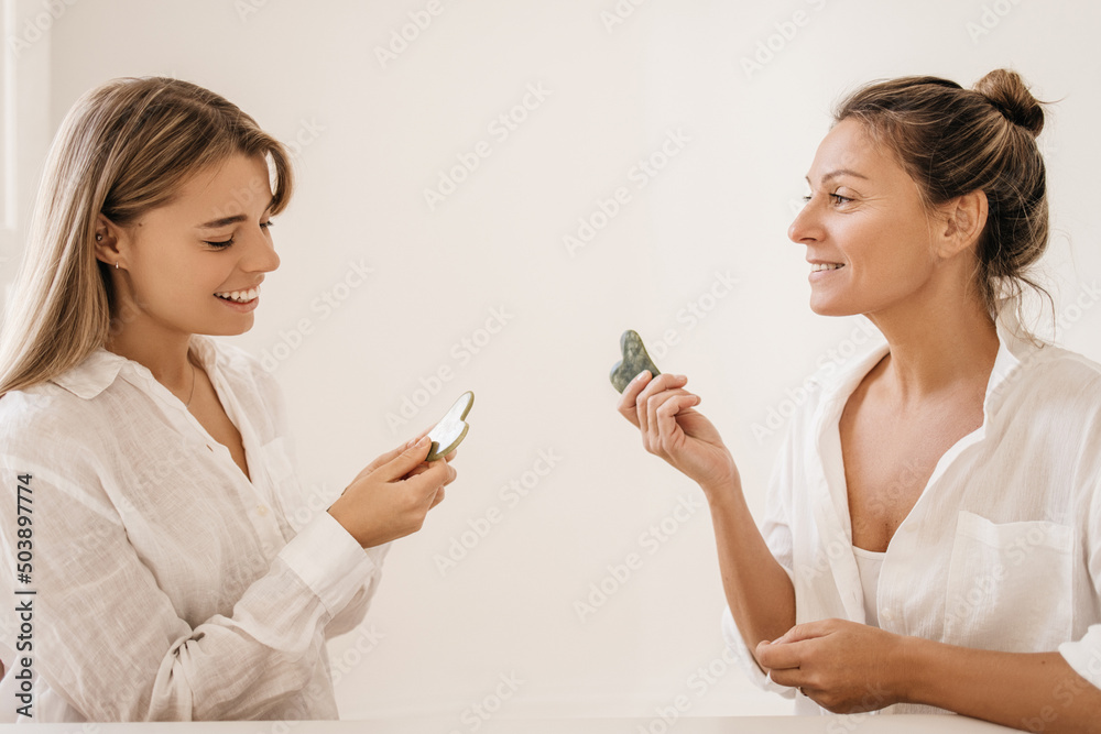 Two pretty caucasian young woman hold gouache scrapers in their hands on white background. Blondes with smiles on their faces wear shirts. Day with skin care and health. Two pretty caucasian young woman hold gouache scrapers in their hands on white background. Blondes with smiles on their faces wear shirts. Day with skin care and health.