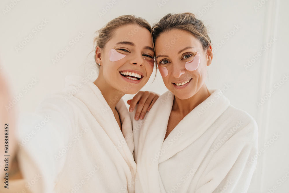 Happy caucasian mother with daughter wearing hydrogel patches smiling looking at camera on white background. Blondes wear robes to take care of their skin. Cosmetic natural trend. - obrazy, fototapety, plakaty 