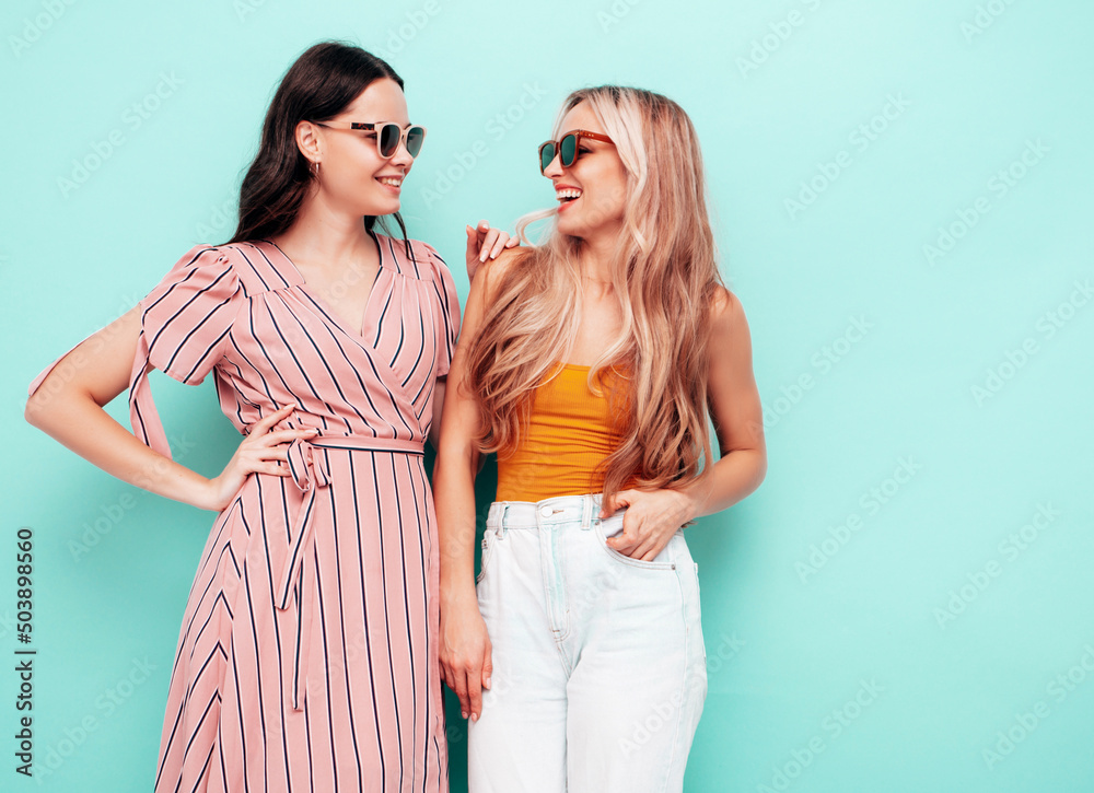 Two young beautiful smiling brunette hipster female in trendy summer clothes. carefree women posing near blue wall. Positive models having fun. Cheerful and happy