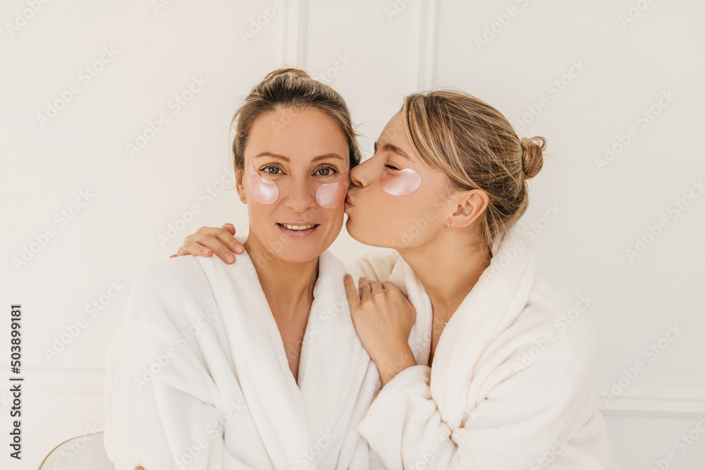 Cute young caucasian girl kisses her mother on cheek, blonde women in white bathrobes use anti-wrinkle patches indoors. Rituals of female beauty - obrazy, fototapety, plakaty 