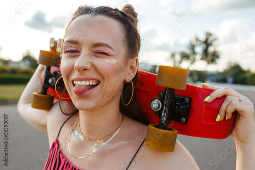 Positive young teenage female skater with skateboard showing tongue and making funny face while looking at camera in city street against sunset sky in summer  photo