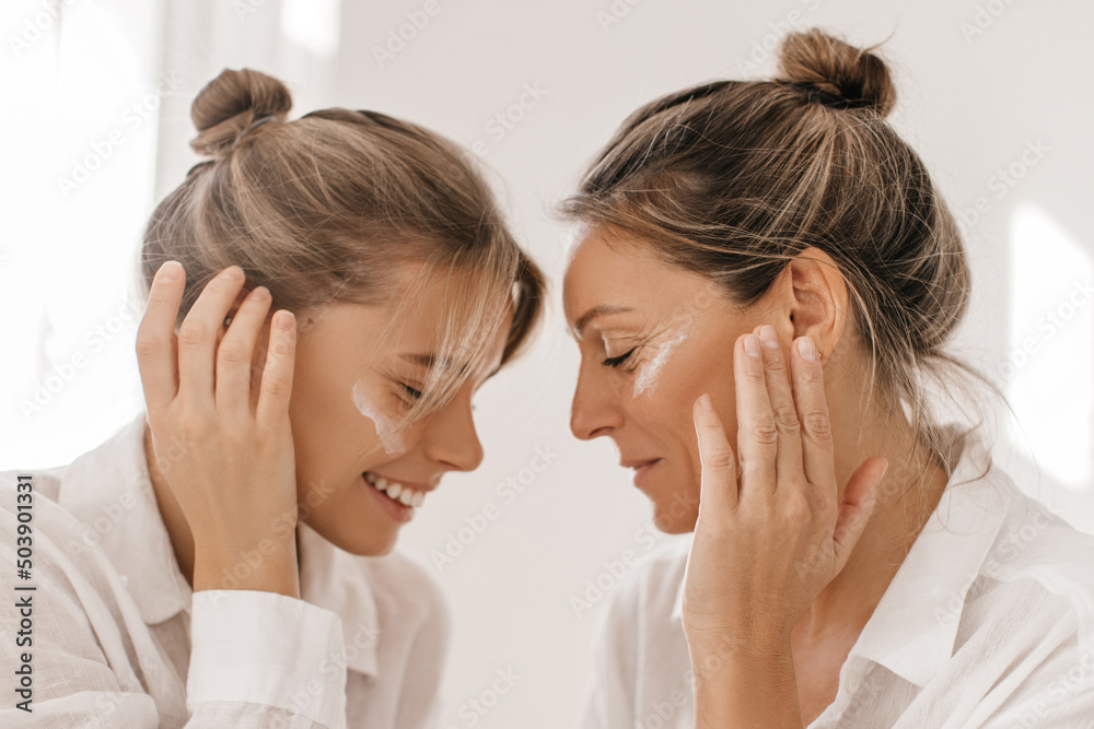 Close-up in profile of two caucasian ladies of different ages with closed eyes on white background. Blonde girls with bun on their heads in shirts. Day with skin care and health.