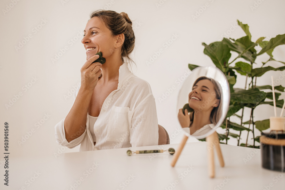Pretty fair-skinned young blonde move special gouache scraper across their face sitting in studio. Women wear white shirts in good mood are doing massage. Skin care and health concept Pretty fair-skinned young blonde move special gouache scraper across their face sitting in studio. Women wear white shirts in good mood are doing massage. Skin care and health concept