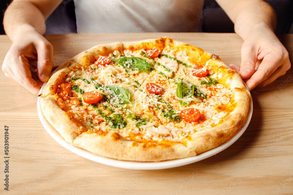 Italian pizza with chicken, cherry tomatoes, mozzarella and parmesan cheese, basil on plate on wooden table and male caucasian hands. Top front view, lifestyle photo, closeup.