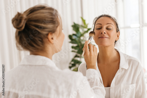 Pretty adult caucasian woman uses services of make-up artist using powder in light room. Blonde woman in white shirt is preparing for meeting. Natural beauty, skin care concept