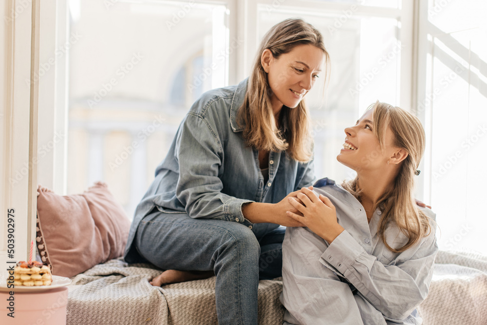 Two beautiful caucasian sisters of different ages are smiling looking at each other sitting by window during day. Blondes wear shirts and jeans. Love expression concept Two beautiful caucasian sisters of different ages are smiling looking at each other sitting by window during day. Blondes wear shirts and jeans. Love expression concept