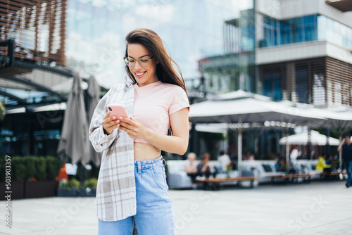 Portrait of cheerful fit female in trendy wear spending time on street using smartphone, beautiful millennial hipster girl blogger holding mobile phone